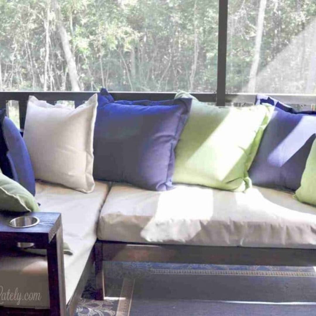 A DIY outdoor sectional sofa on a screened-in porch, featuring light grey cushions and an assortment of blue and green throw pillows.