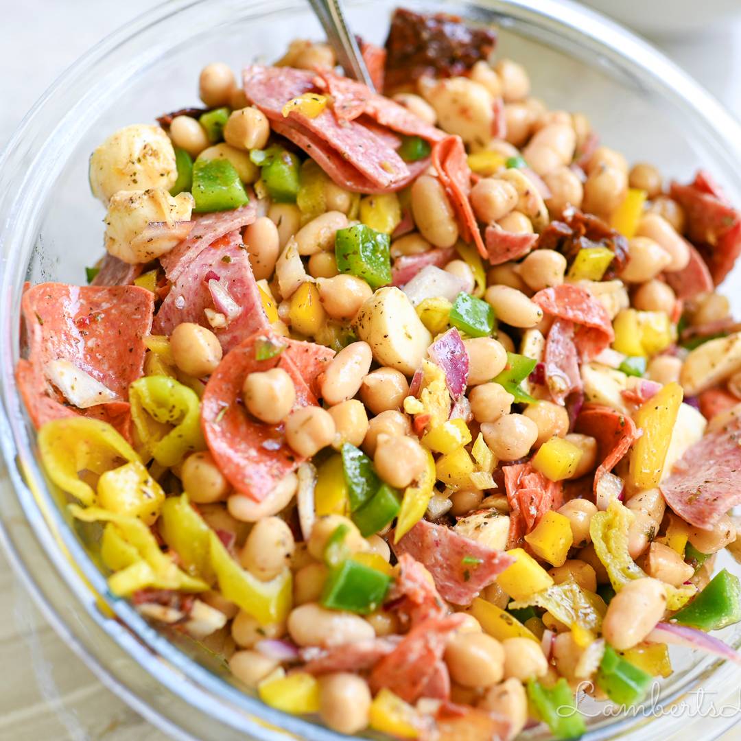 A glass bowl filled with a colorful Mediterranean-style bean salad featuring white beans, sliced salami, green and yellow bell peppers, and red onion in a light vinaigrette.