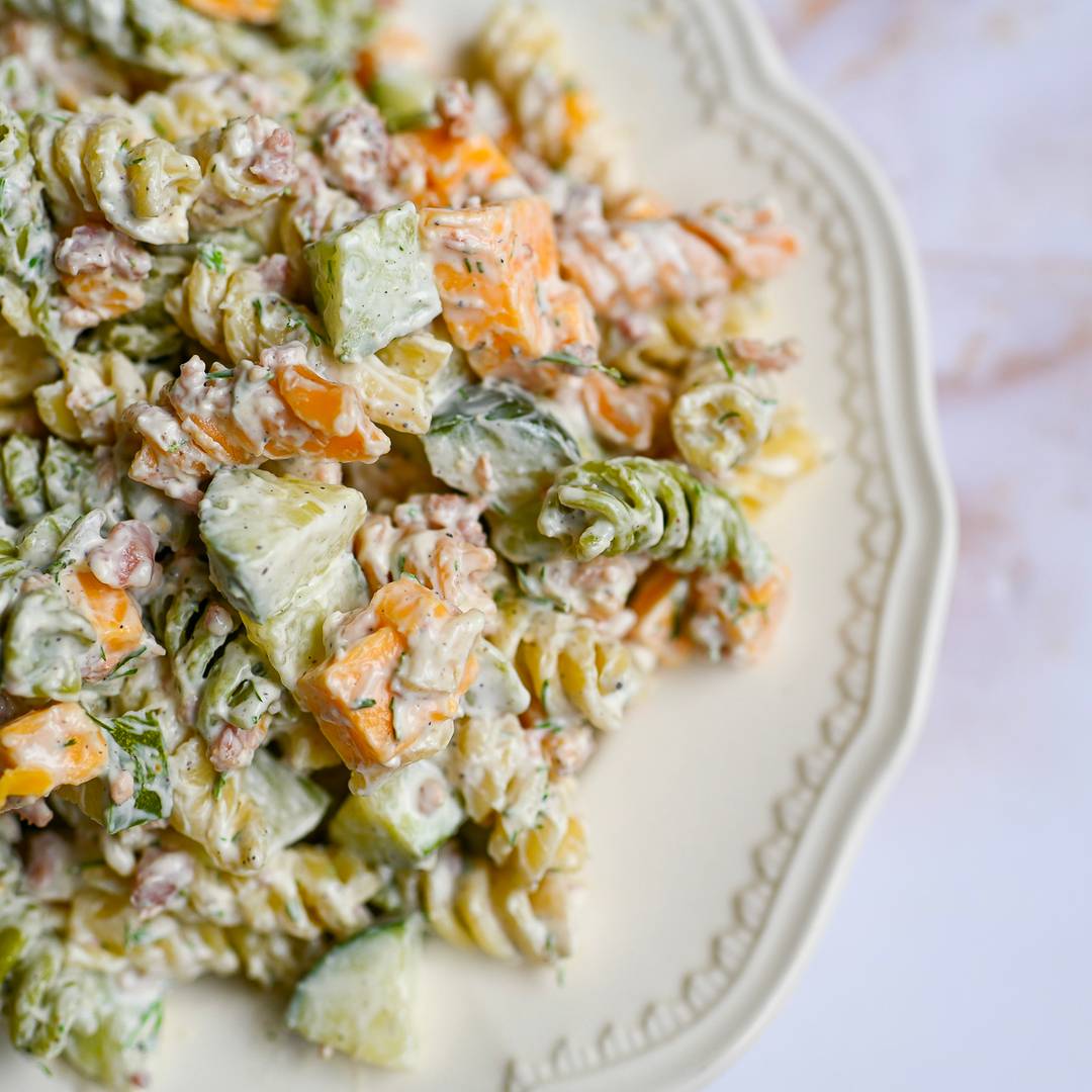 A close-up view of a creamy pasta salad served on a decorative white plate, featuring rotini noodles, diced cucumbers, cheese cubes, bacon bits, and fresh dill.