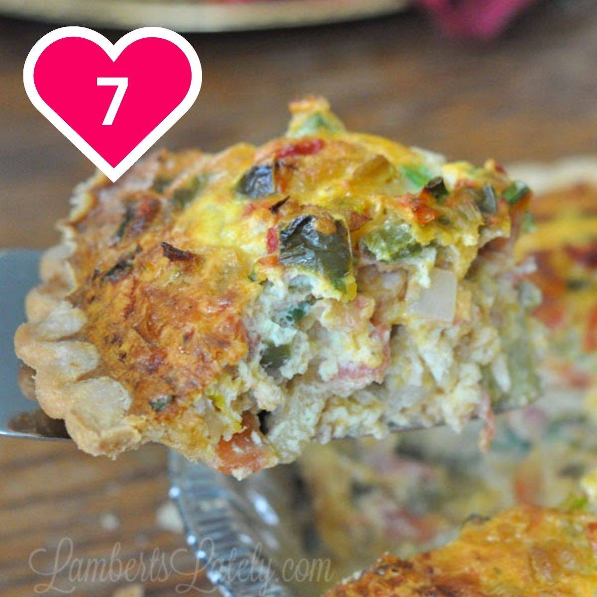 A metal spatula lifting a slice of a savory quiche out of a glass pie dish. The quiche has a crimped pastry crust and a filling loaded with eggs, cheese, green peppers, onions, and ham or bacon. A pink heart icon with the number 7 is in the upper left.