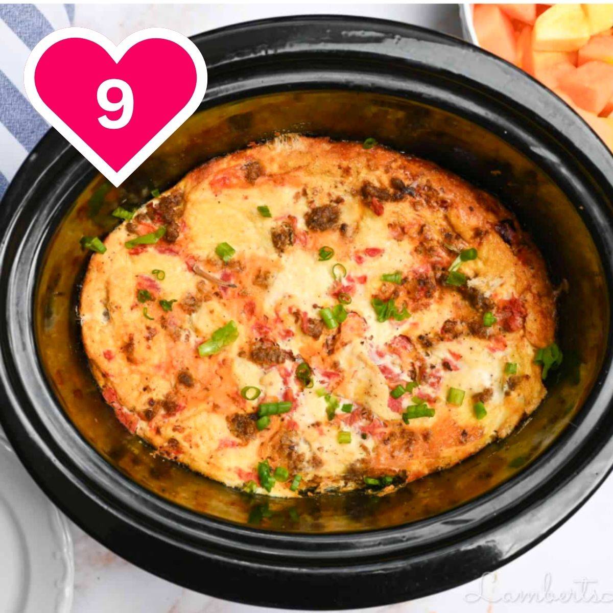An overhead view of a savory breakfast casserole or frittata cooked inside a black oval slow cooker. The dish is golden-brown with visible bits of melted cheese, sausage or bacon, tomatoes, and a garnish of freshly chopped green onions. A pink heart icon with the number 9 is in the upper left.