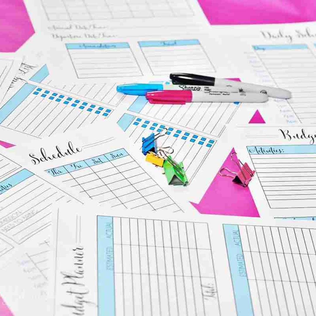 A flat lay of printed travel planning pages including a schedule, budget planner, and activity log. The pages are accented with light blue headers and are scattered on a pink surface with colorful binder clips and Sharpie markers.