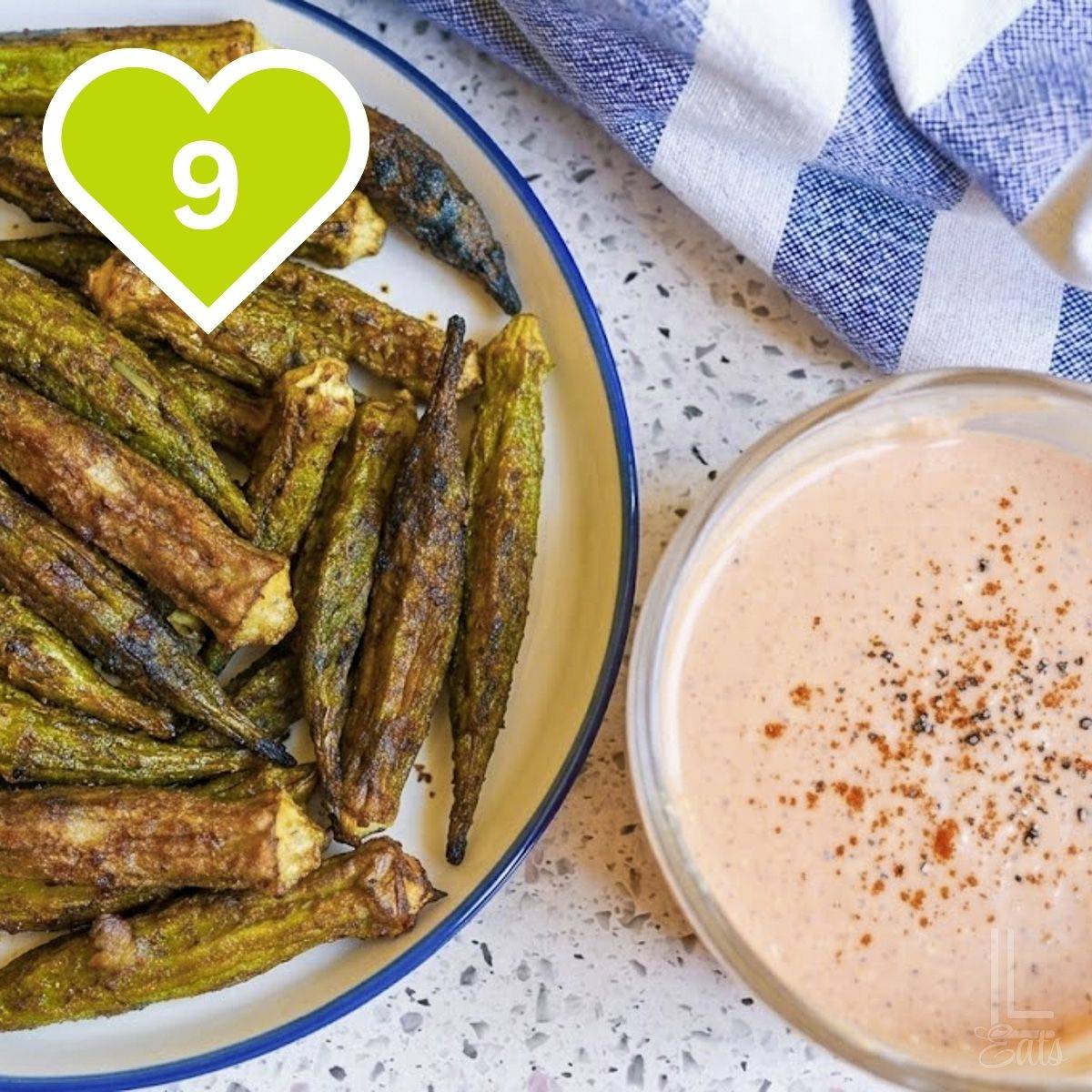 Air fryer roasted okra spears in a shallow glass dish alongside a bowl of creamy dipping sauce.