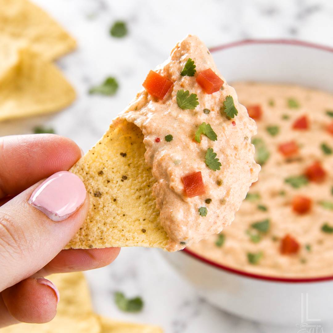 Tortilla chip topped with creamy salsa dip, diced tomatoes, and cilantro, with a bowl of dip in the background.