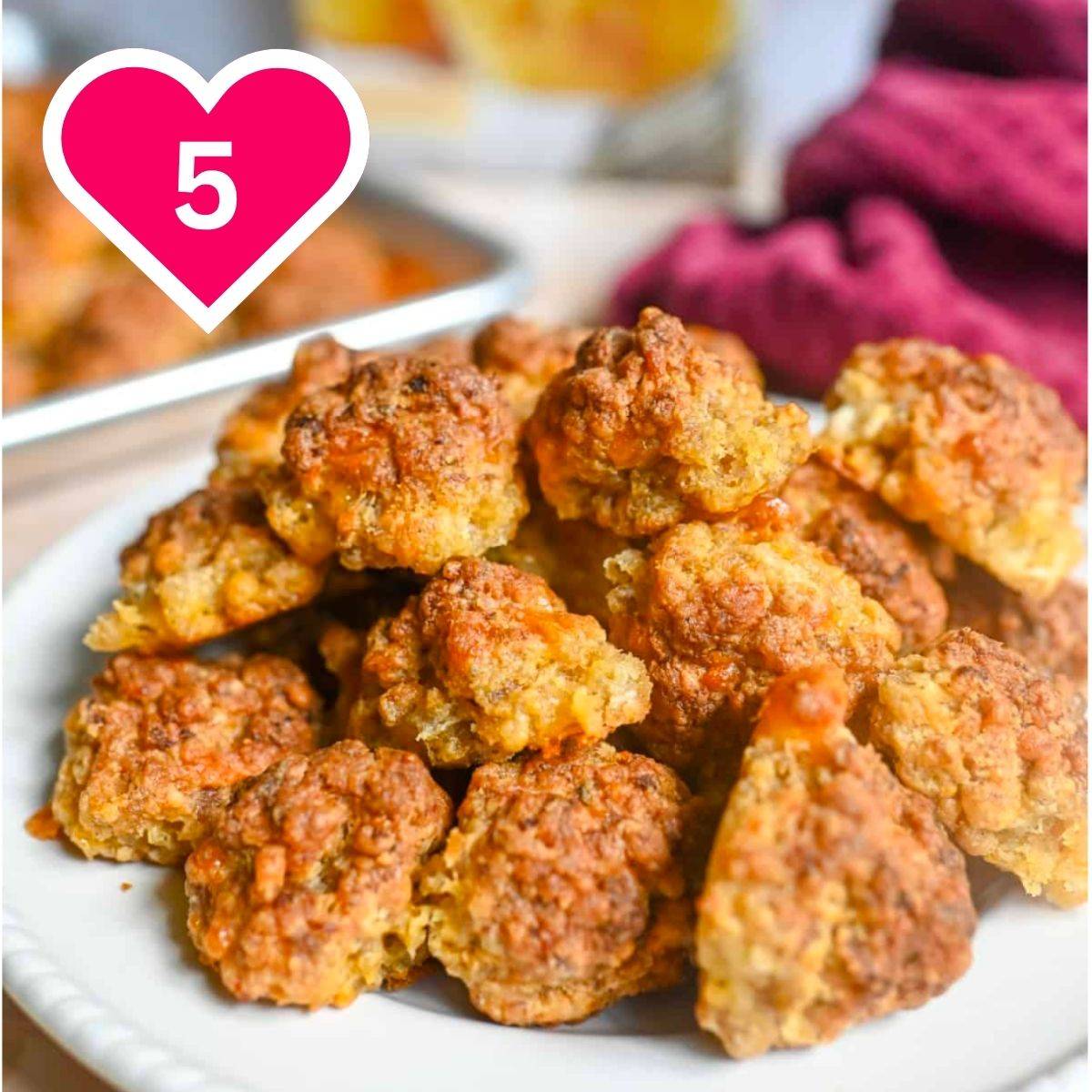 A white plate stacked high with golden-brown, crispy baked sausage balls. In the background, a metal baking sheet and a dark red kitchen towel are softly blurred. A pink heart icon with the number 5 is in the upper left.