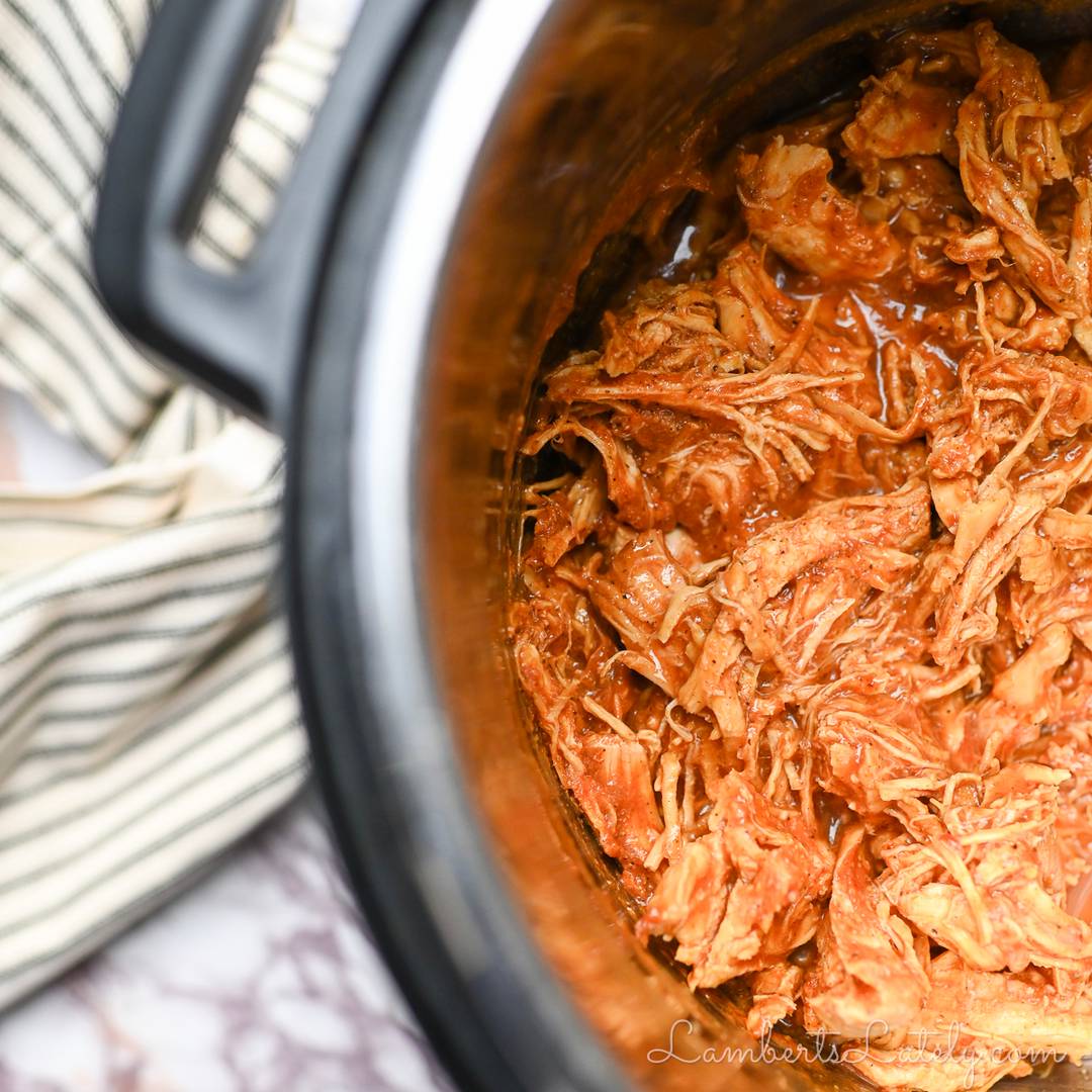 A close-up, overhead view of shredded salsa chicken or buffalo chicken inside a stainless steel Instant Pot insert, showing a rich, red sauce and tender meat.