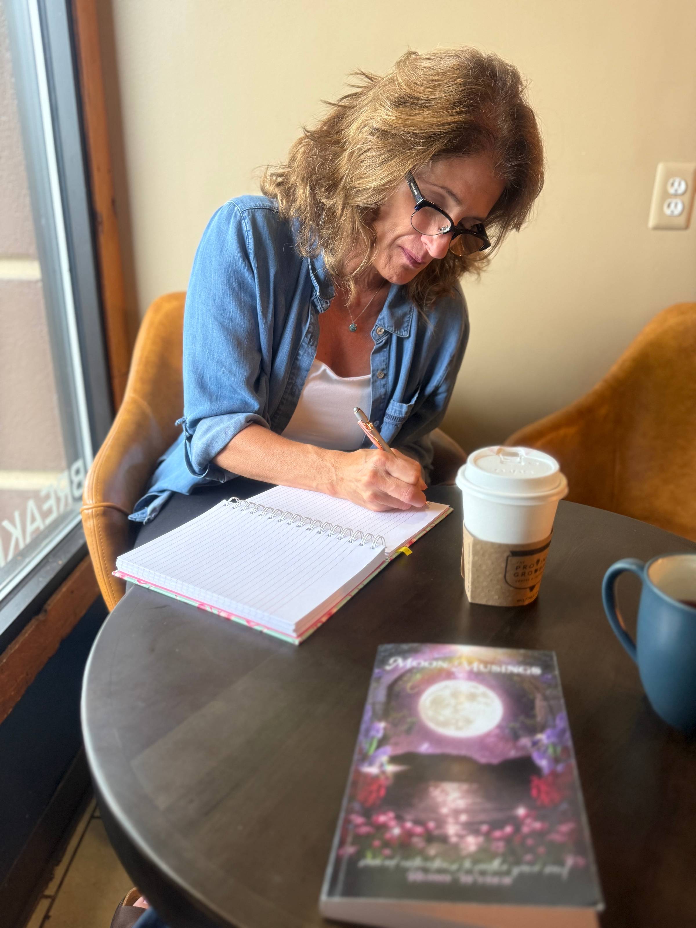 Leslie writing in a notebook at a coffee shop with a cup of tea beside her