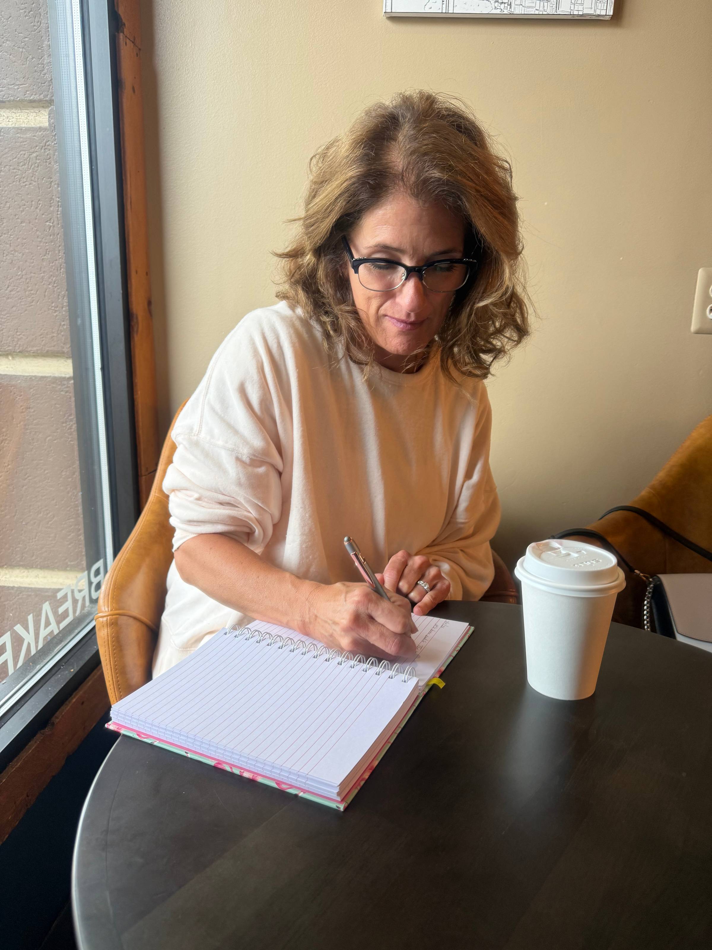 Leslie writing in a notebook at a coffee shop with a cup of tea beside her