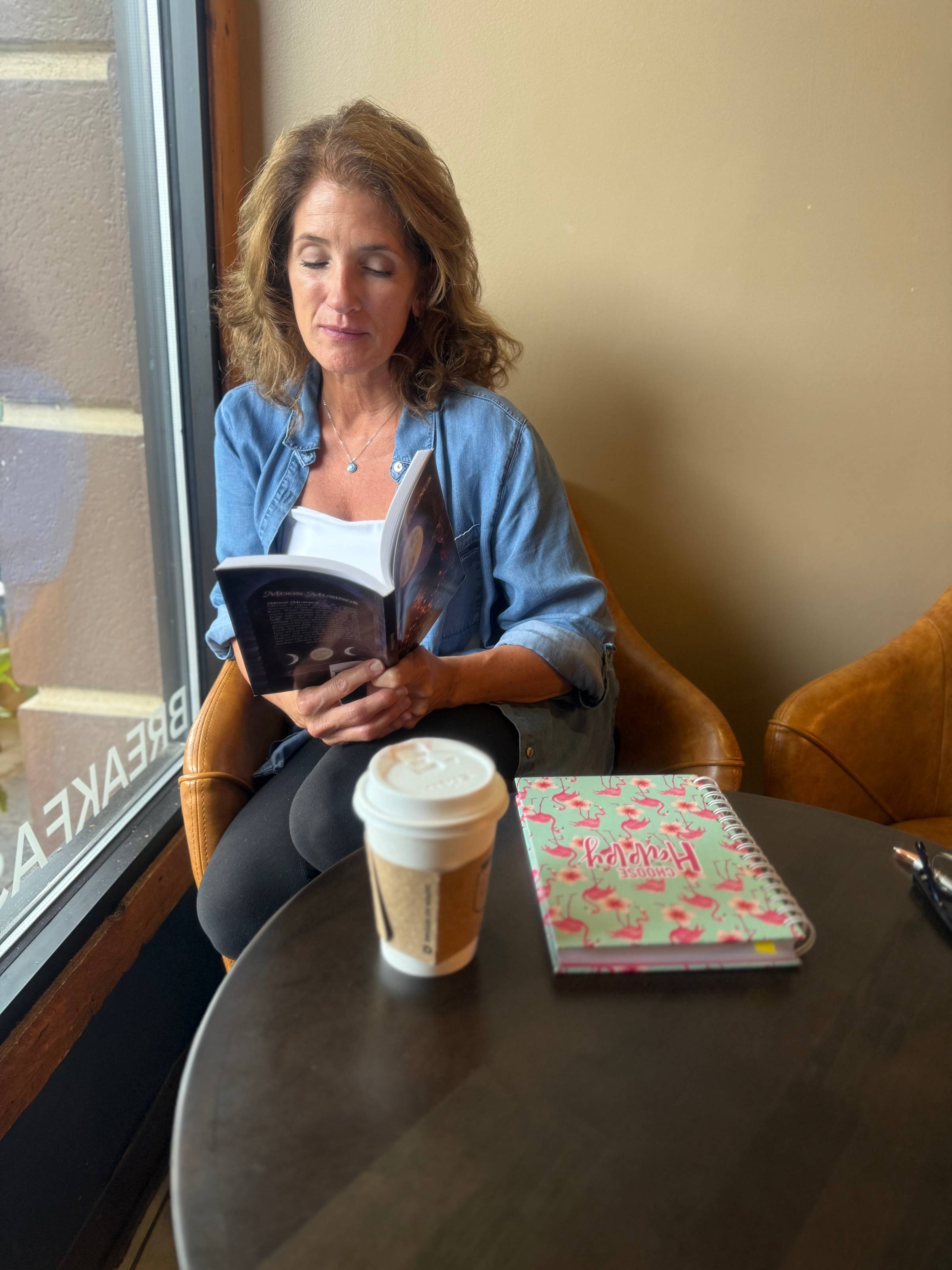 Leslie reading a book at a coffee shop with a notebook and cooffee on the table.