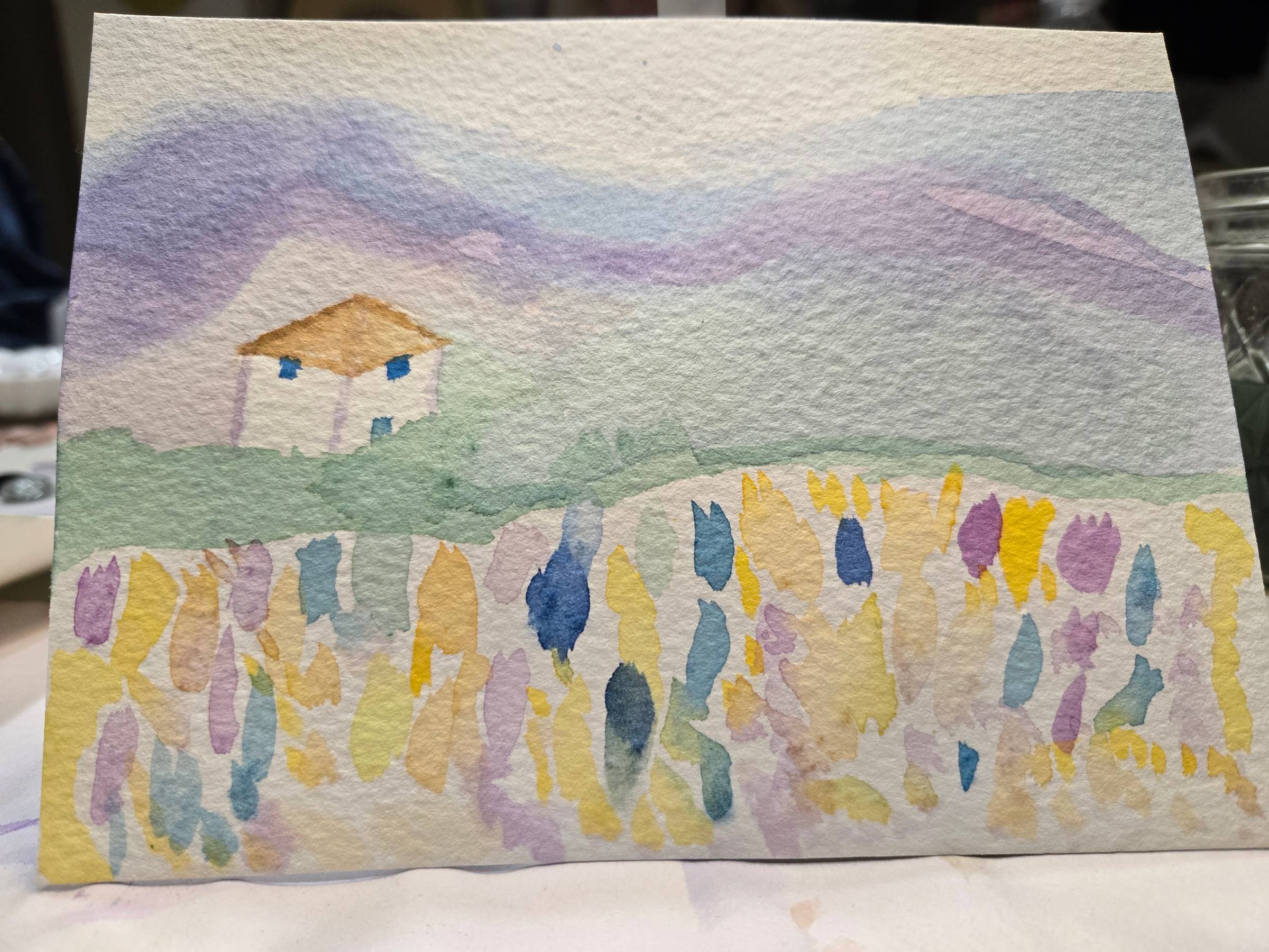 A closeup of a rudimentary watercolor landscape with a white house and abstract flowers in the foreground.