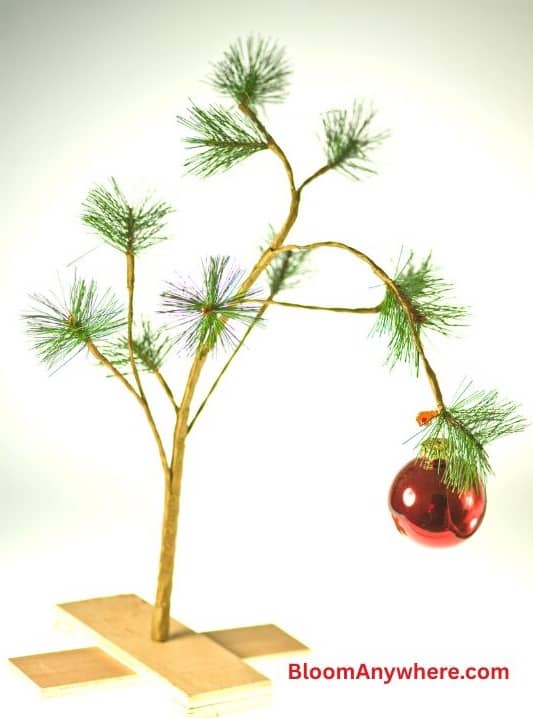 An evergreen sprig anchored to two pieces of wood decorated with a red ornament.