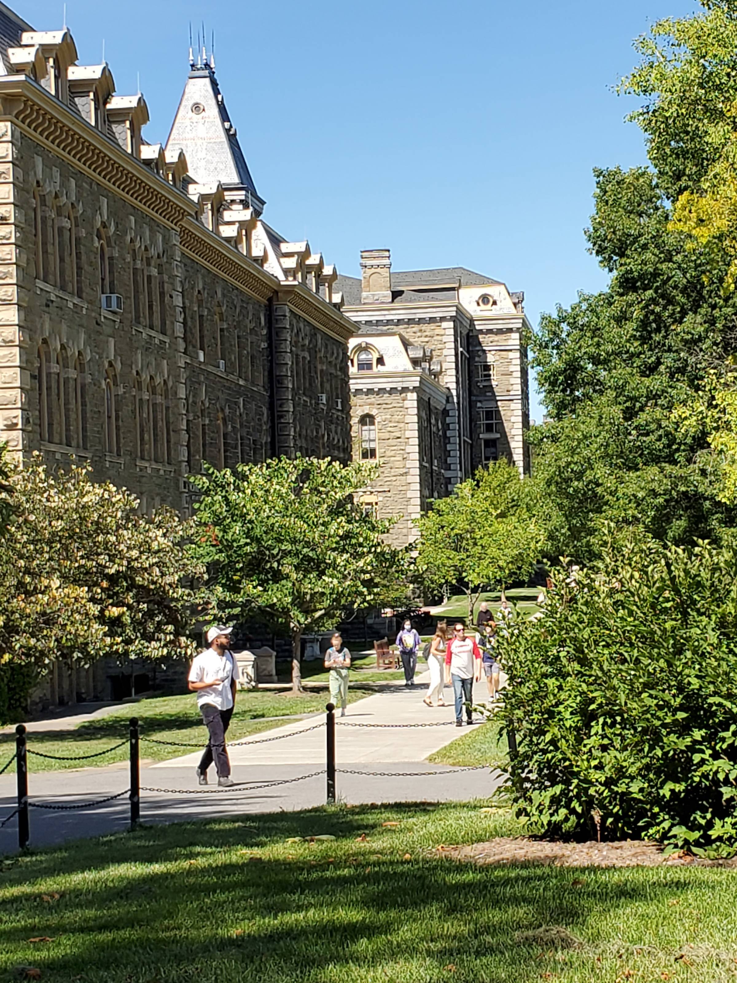 Students walking on a university campus with stone buildings and greenery around them.
