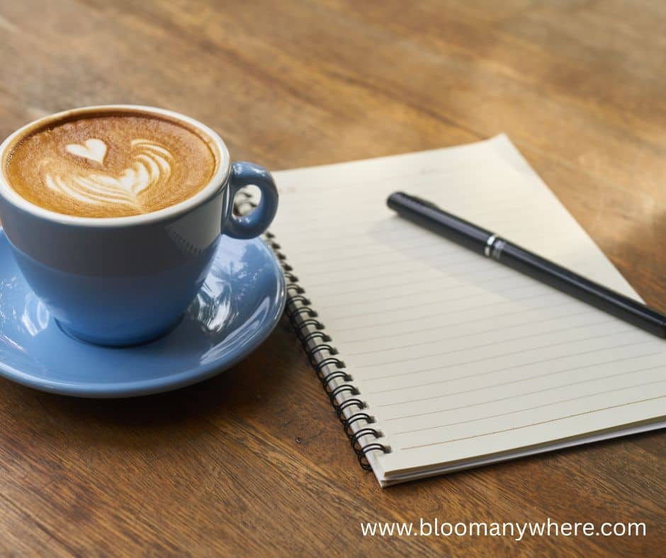 A blank spiral notebook with a black pen on top. Next to the notebook is a blue cup and saucer filled with coffee and topped with foam art in the shape of a heart.