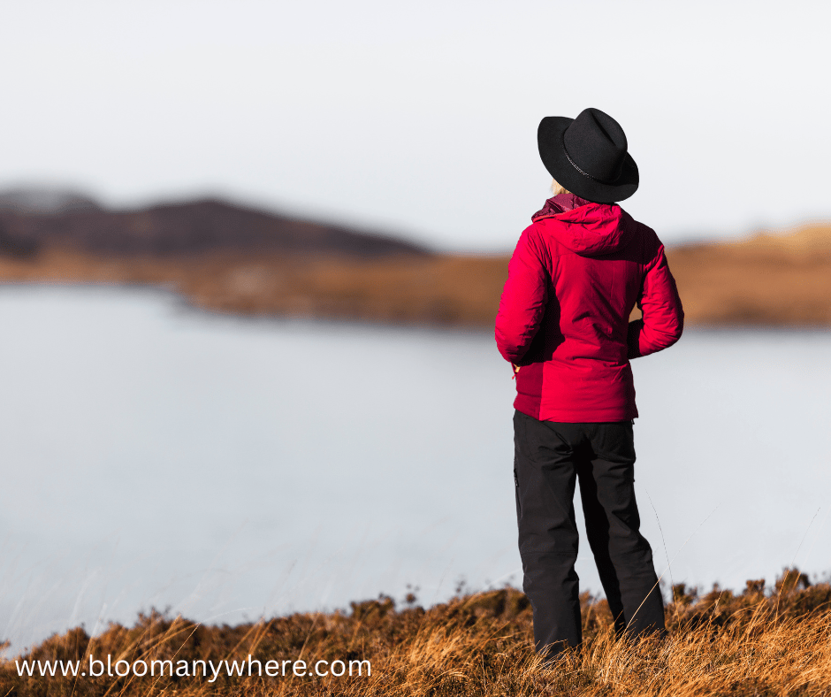 A woman in a red jacket, hat, and black trousers looking across a lake.