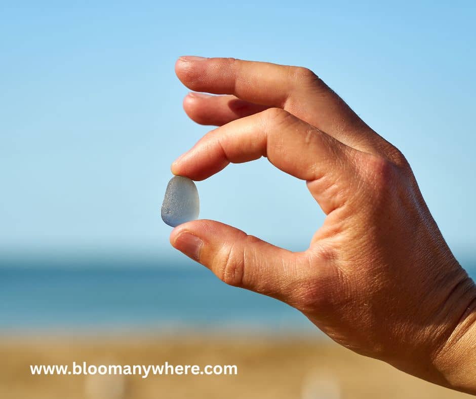 A hand holding a piece of sea glass between the thumb and forefinger with the beach and ocean in the background.