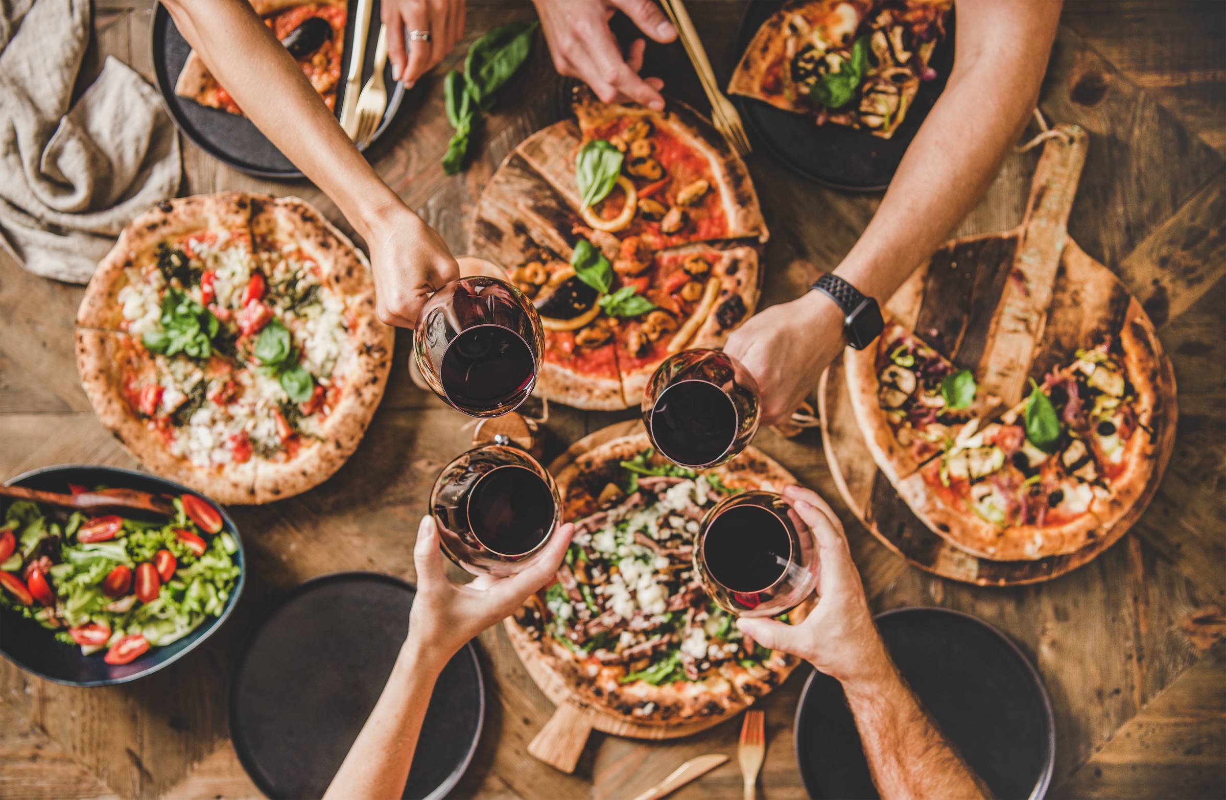 Arms extended over several small pizzas. They hold glasses of red wine in a toast.