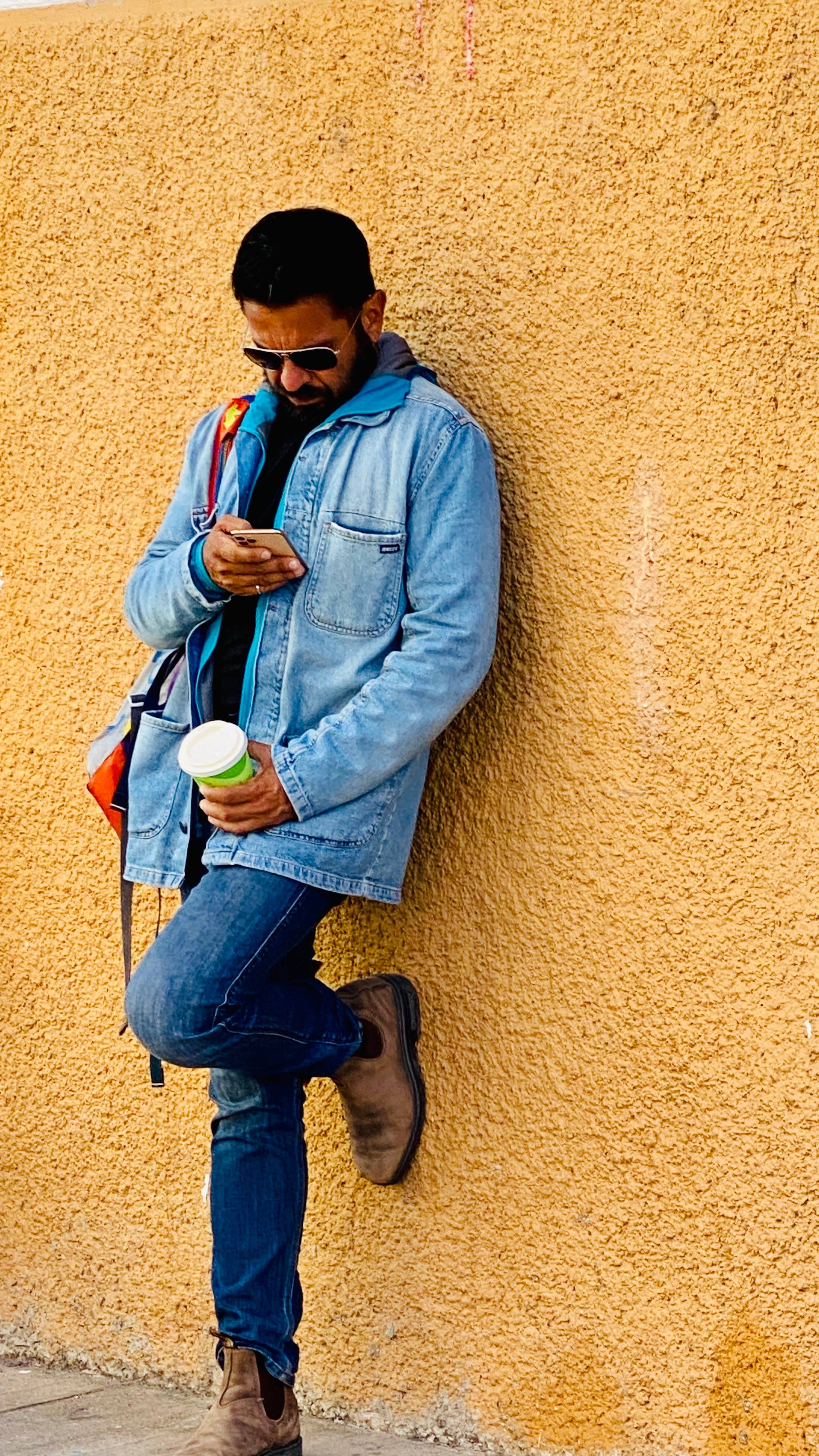 Zaheer leans against a yellow wall, eyes fixed on his phone, coffee in hand — a quiet moment of scrolling that mirrors the tension between presence and performance explored in the letter.
