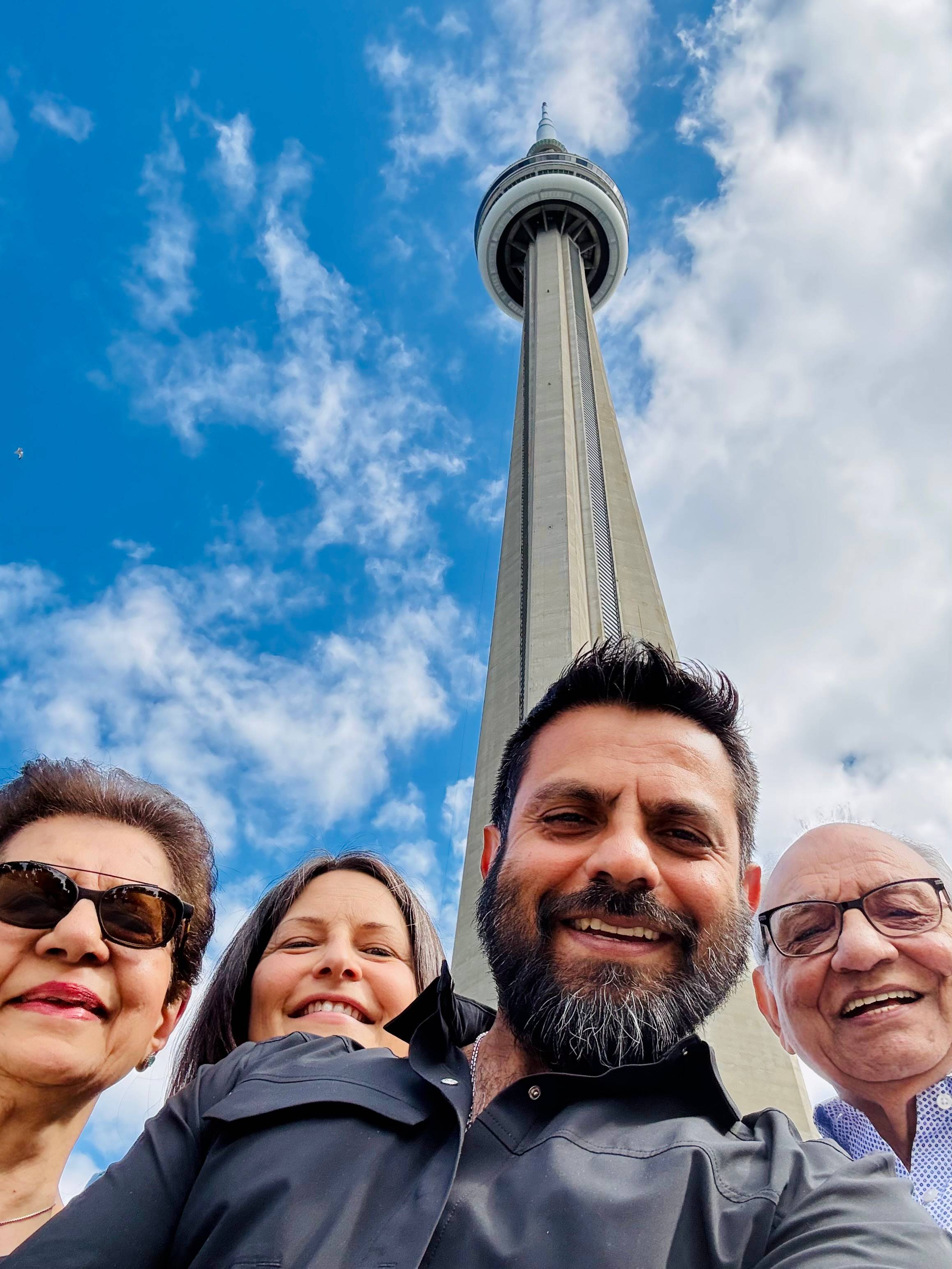 Zaheer standing with his parents and partner beneath the CN Tower in Toronto, all smiling under a bright blue sky.
