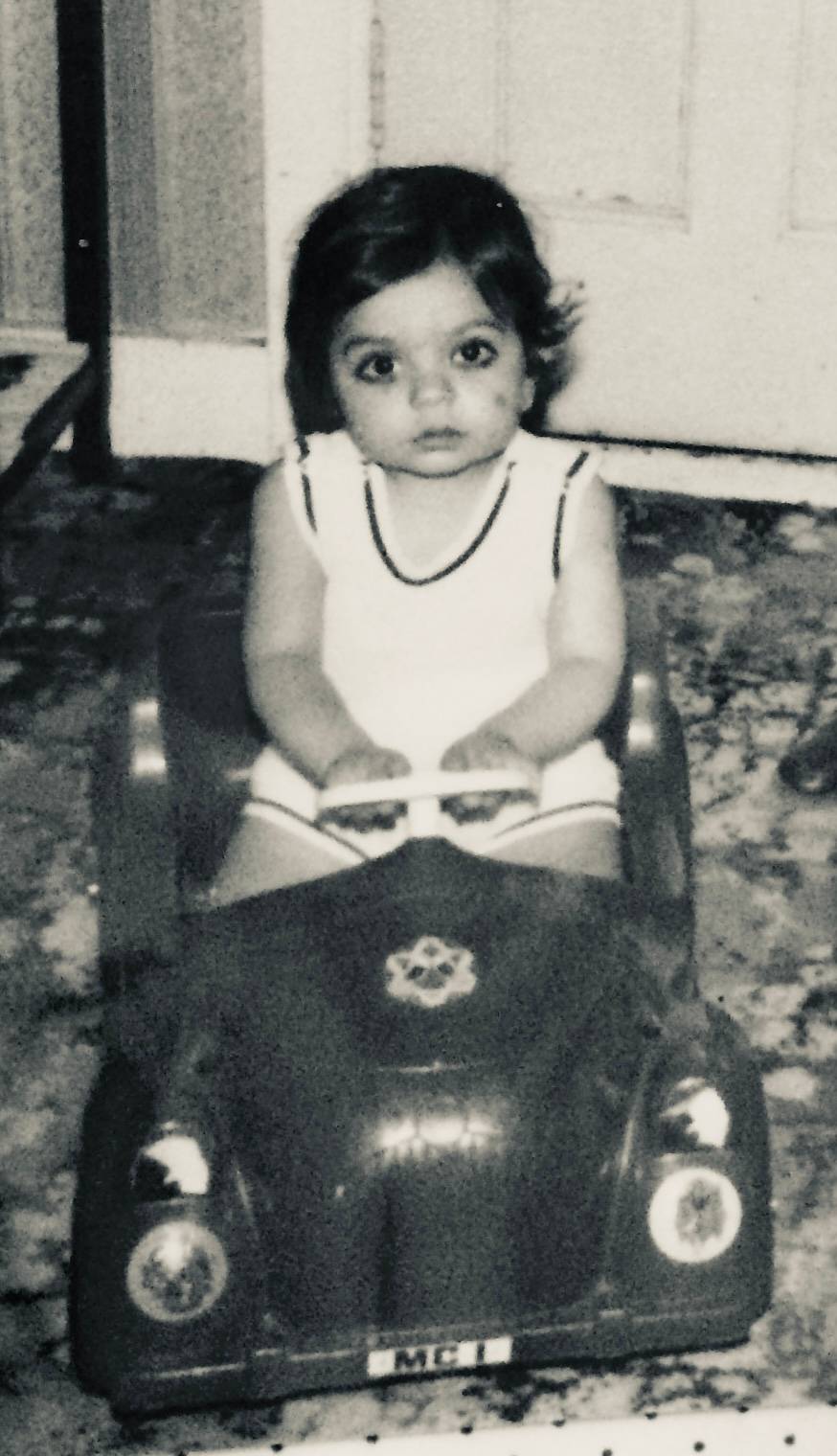Black-and-white photo of a young child sitting still in a toy car, eyes wide with open, unfiltered presence — before language, before self-image.