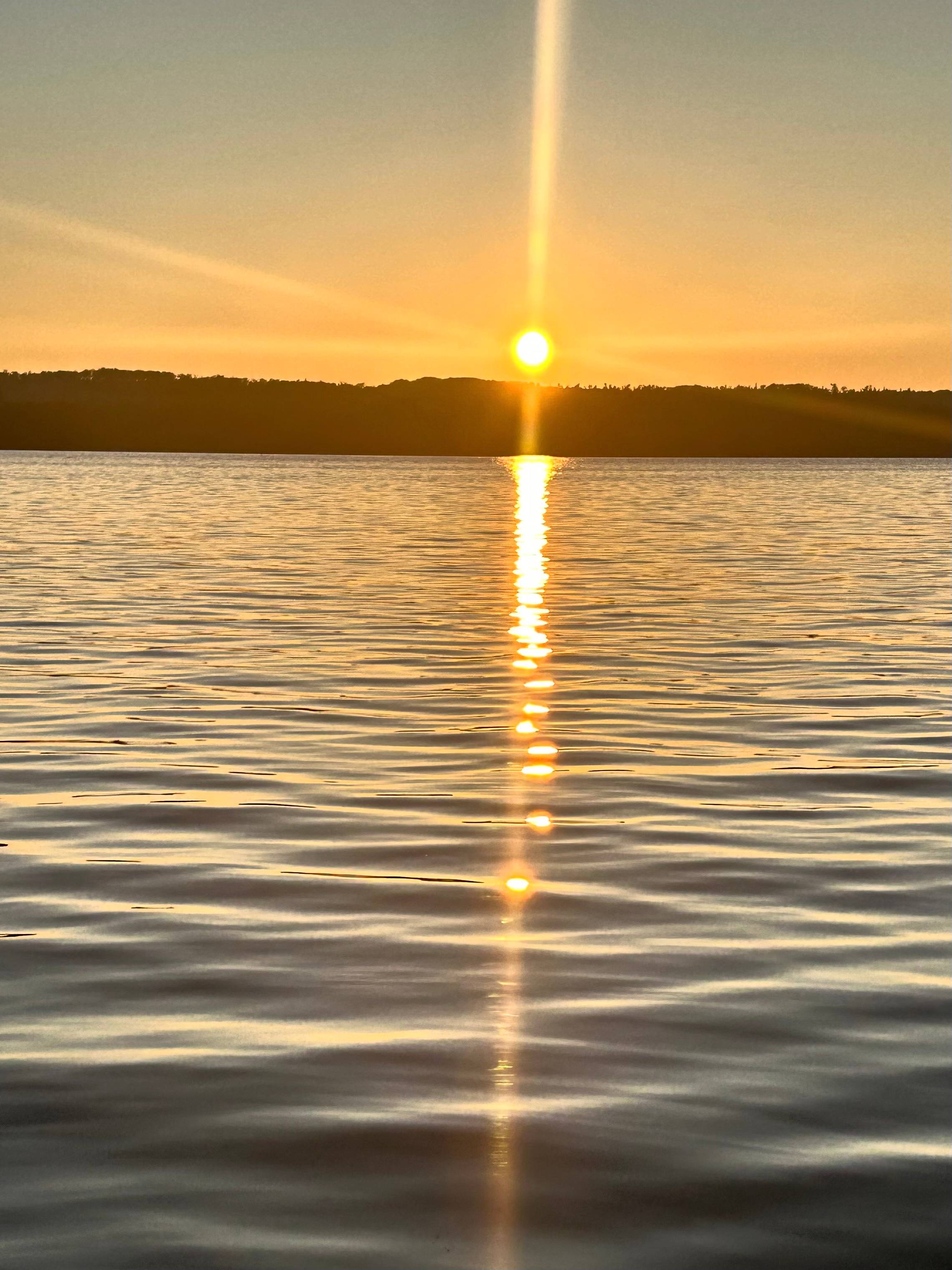 Golden sun sits low over a dark tree-lined horizon, casting a soft vertical reflection across still rippled water. The moment feels suspended — neither beginning nor end — just presence