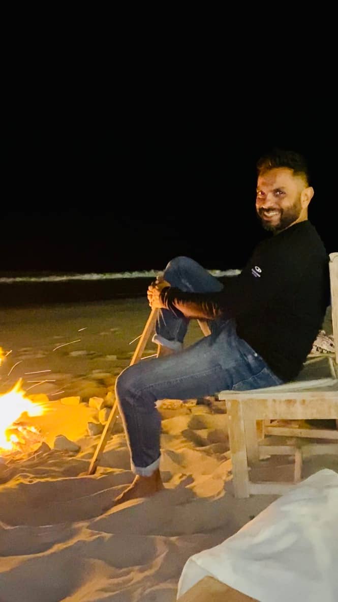 Zaheer sitting barefoot by a beach bonfire at night, smiling gently while gazing sideways. A quiet fire glows in the sand nearby, surrounded by darkness and ocean waves in the background. His posture is relaxed, legs folded, holding a walking stick — at p