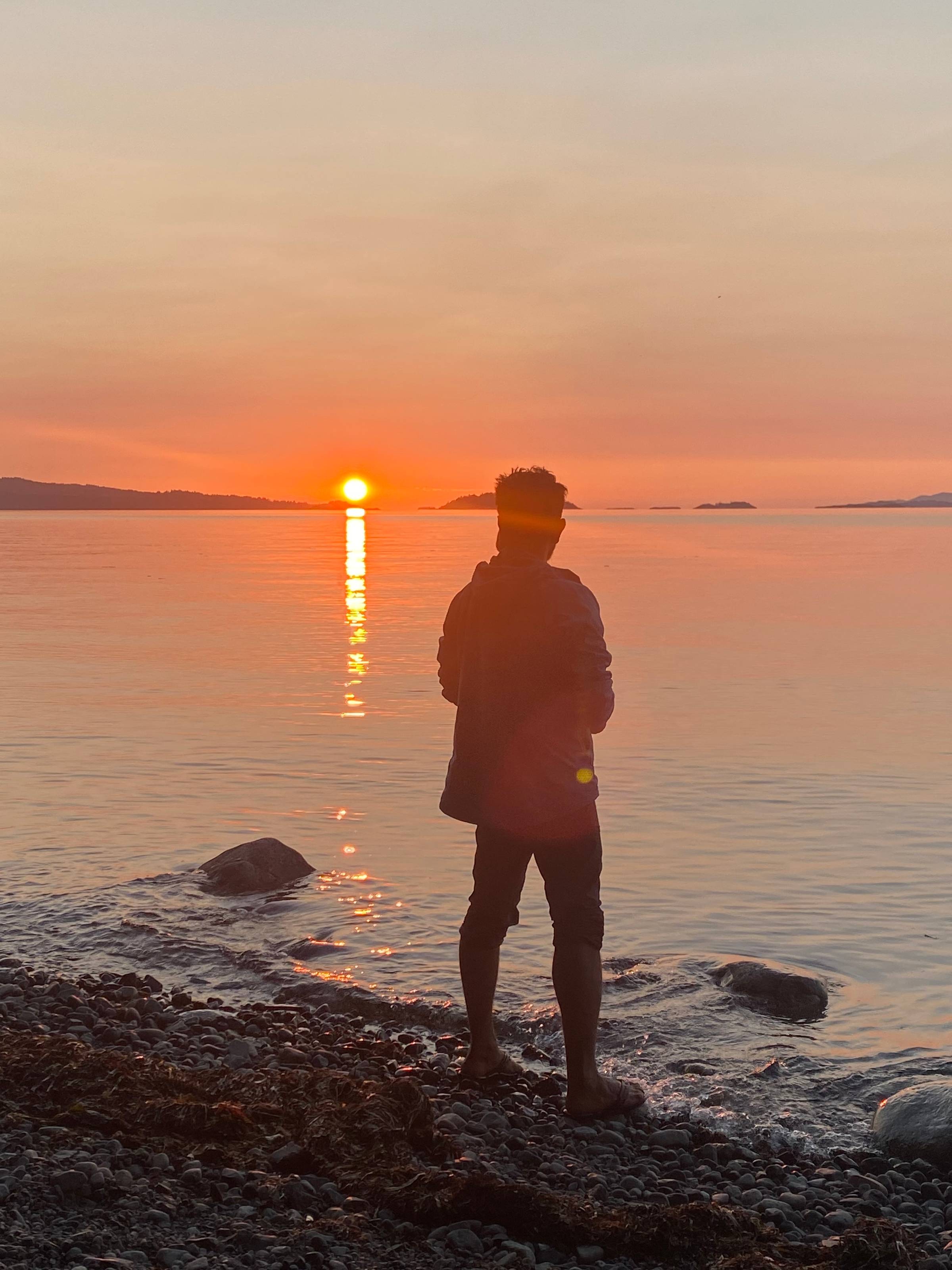 A barefoot Zaheer stands on a rocky shore at sunset, gazing out across a calm ocean. The sun glows near the horizon, casting a golden reflection across the water. The sky is painted in soft hues of orange and pink. His silhouette is peaceful, still, and r