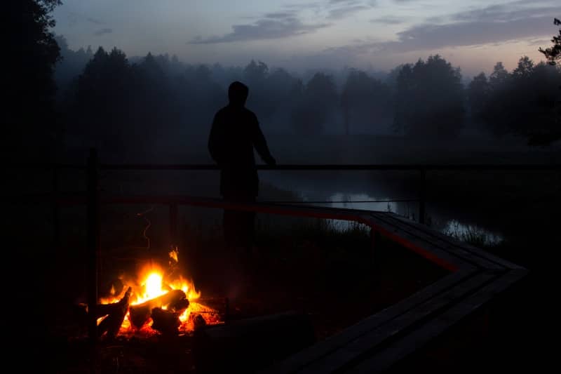 silhouette photo of a person standing near body of water and bonfire