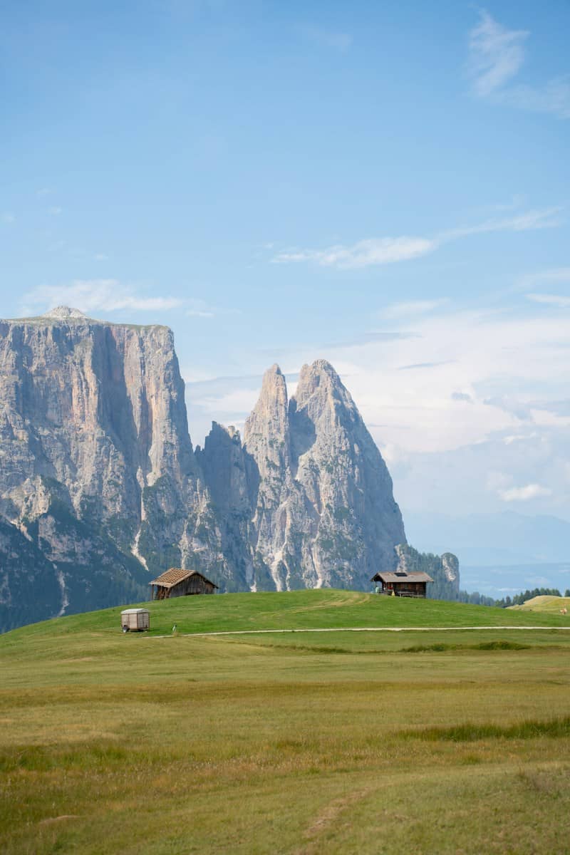 Two wooden huts on a green meadow below the jagged peaks of the Dolomites