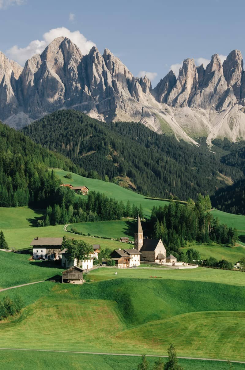 A small village with a church in a green valley below the jagged Odle Mountains