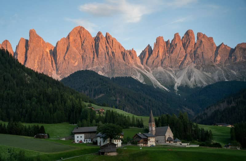A small village with a church in a green valley below jagged mountain peaks