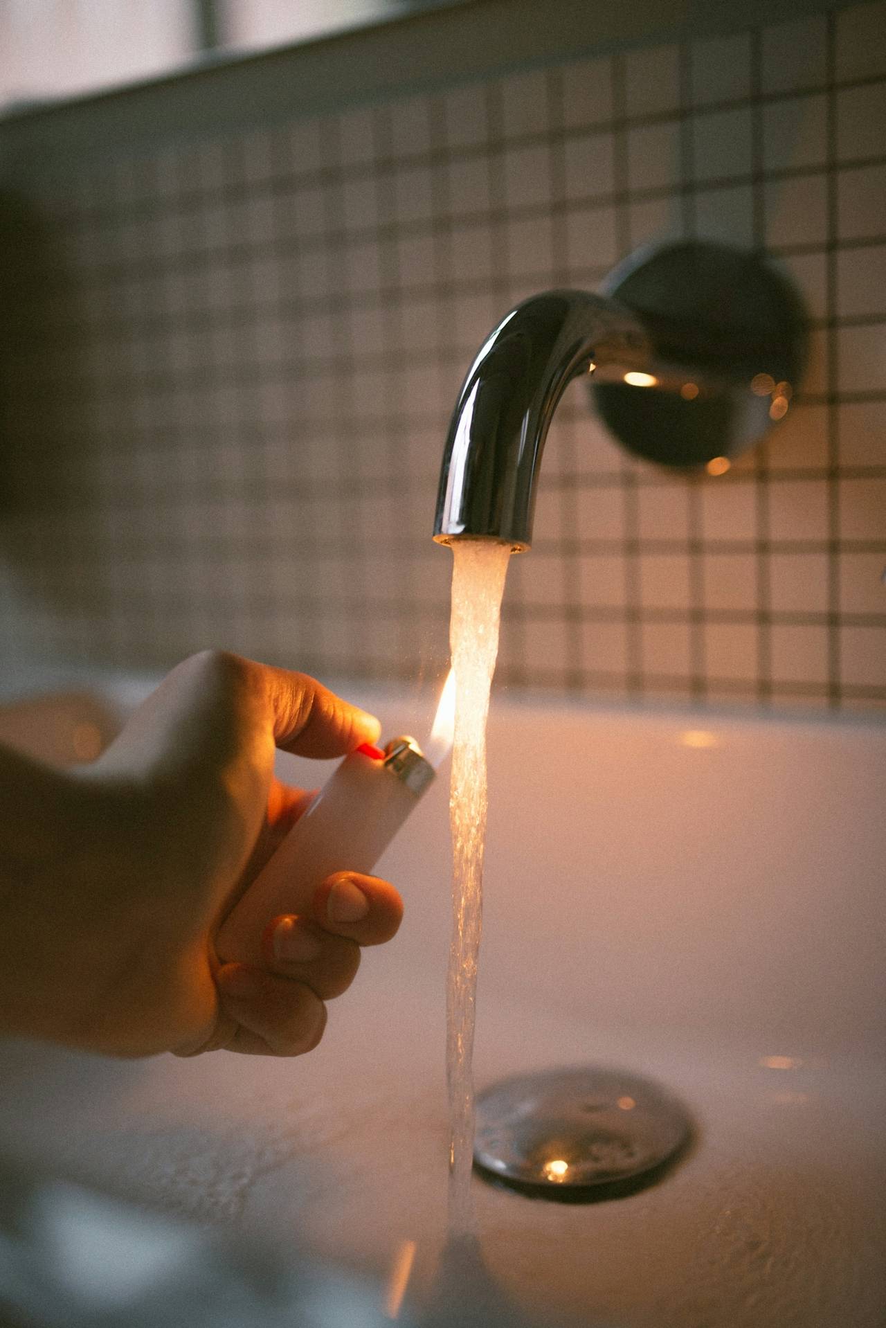 A photo by Chris Abatzis, a stream of water pouring out of a faucet, an a woman's hand holding a lit lighter to it. 