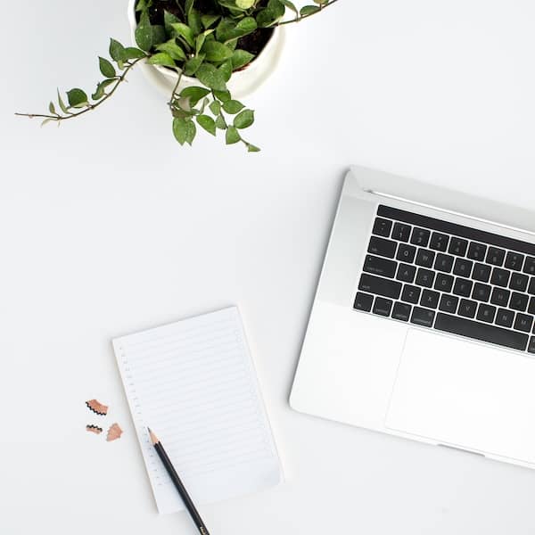 a laptop and notepad sit on a white desk with a pencil and pencil shavings ans plant