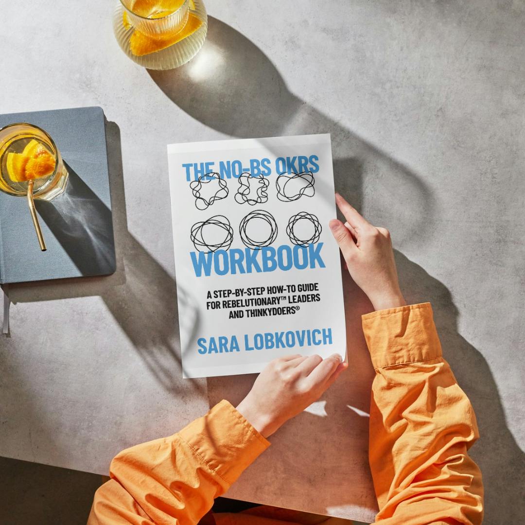 The white book, The No-BS OKRs Workbook is 8.5" by 11" and sits on a slate table, held by a person wearing an orange long-sleeved shirt.