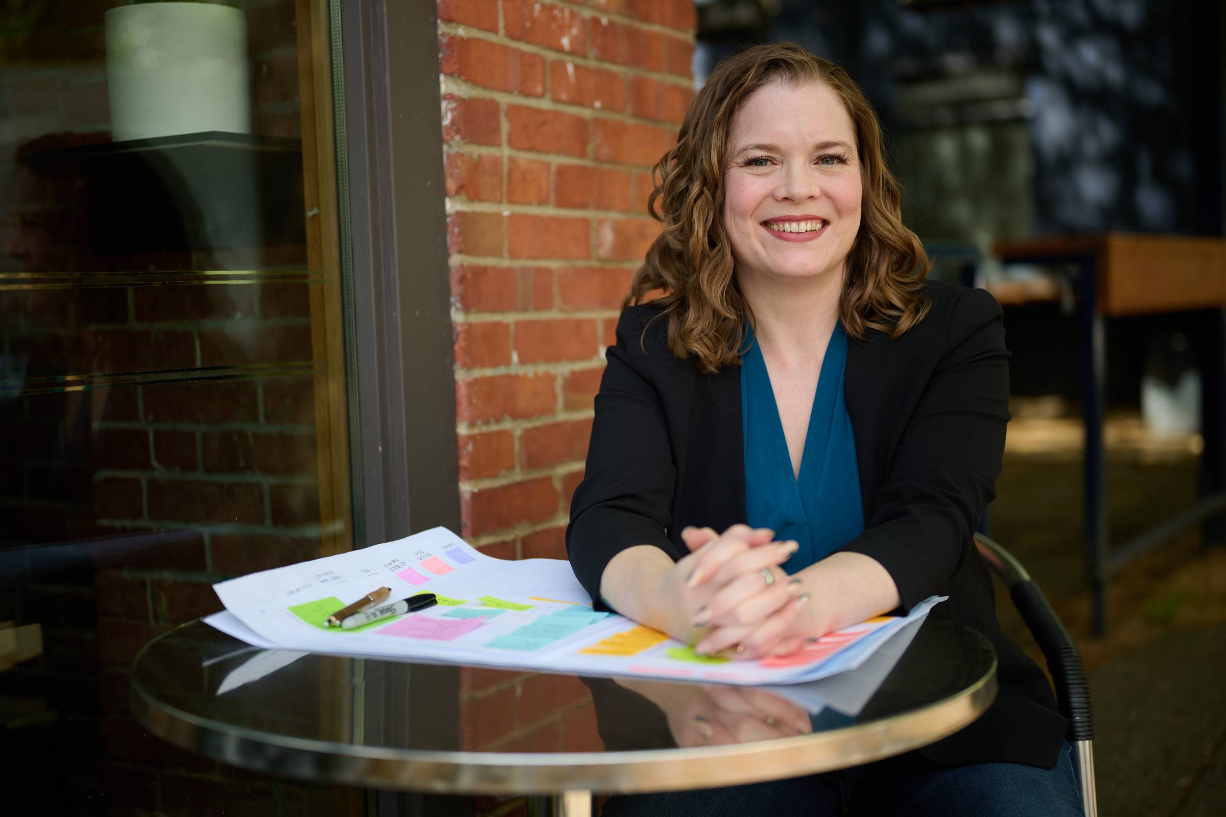 Sara is a white woman with shoulder-length wavy dark blonde hair. She is smiling. She wears a blue shirt under a black jacket. Her arms are resting on a table, with paper covered in sticky notes on it.