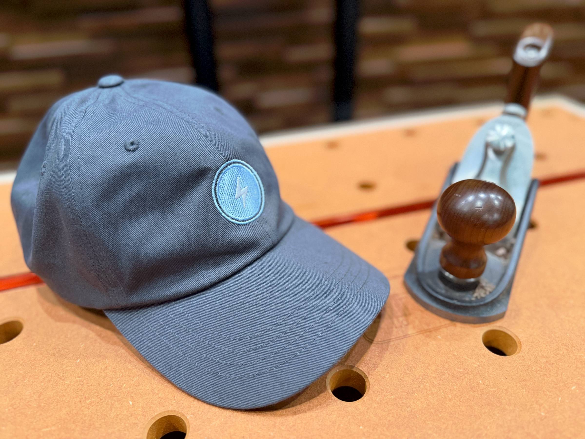Dark blue baseball cap with a light blue lightning bolt logo resting on a woodworking bench beside a metal hand plane with a wooden handle.