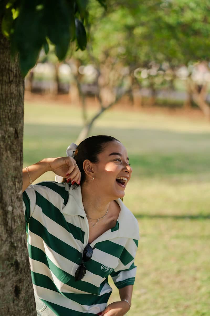 Happy young woman laughing outdoors, leaning against a tree.