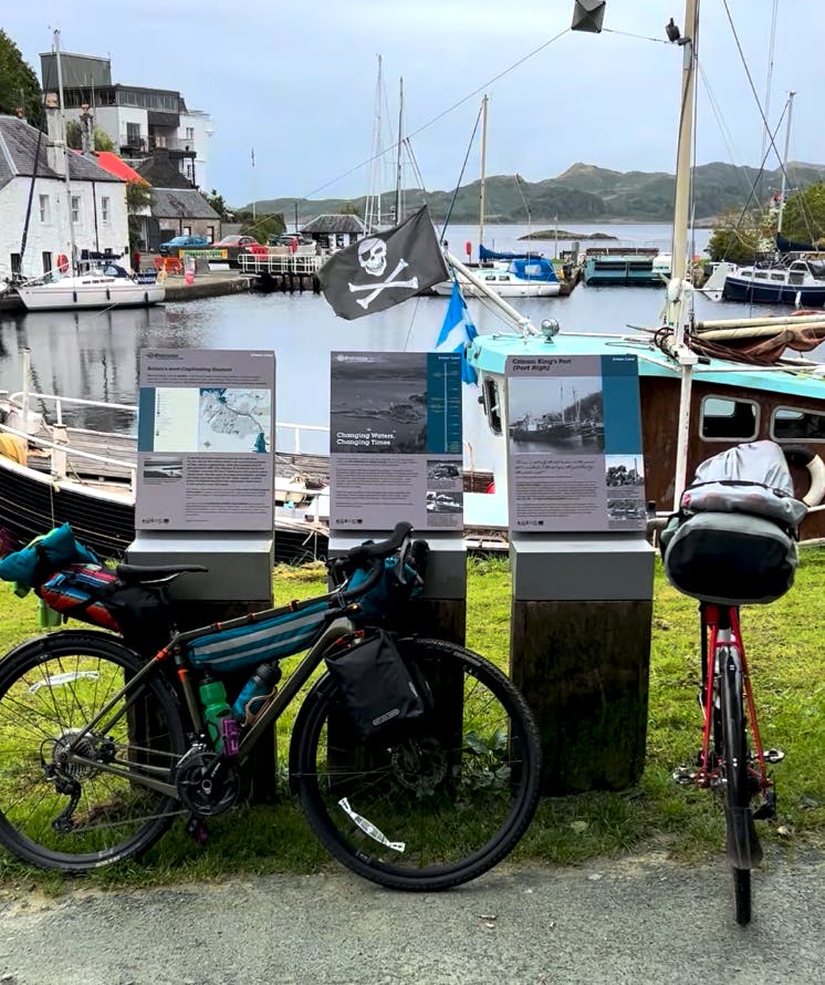 Two touring bikes propped against a road sign in front of a harbor, where the ship behind is flying a pirate flag.