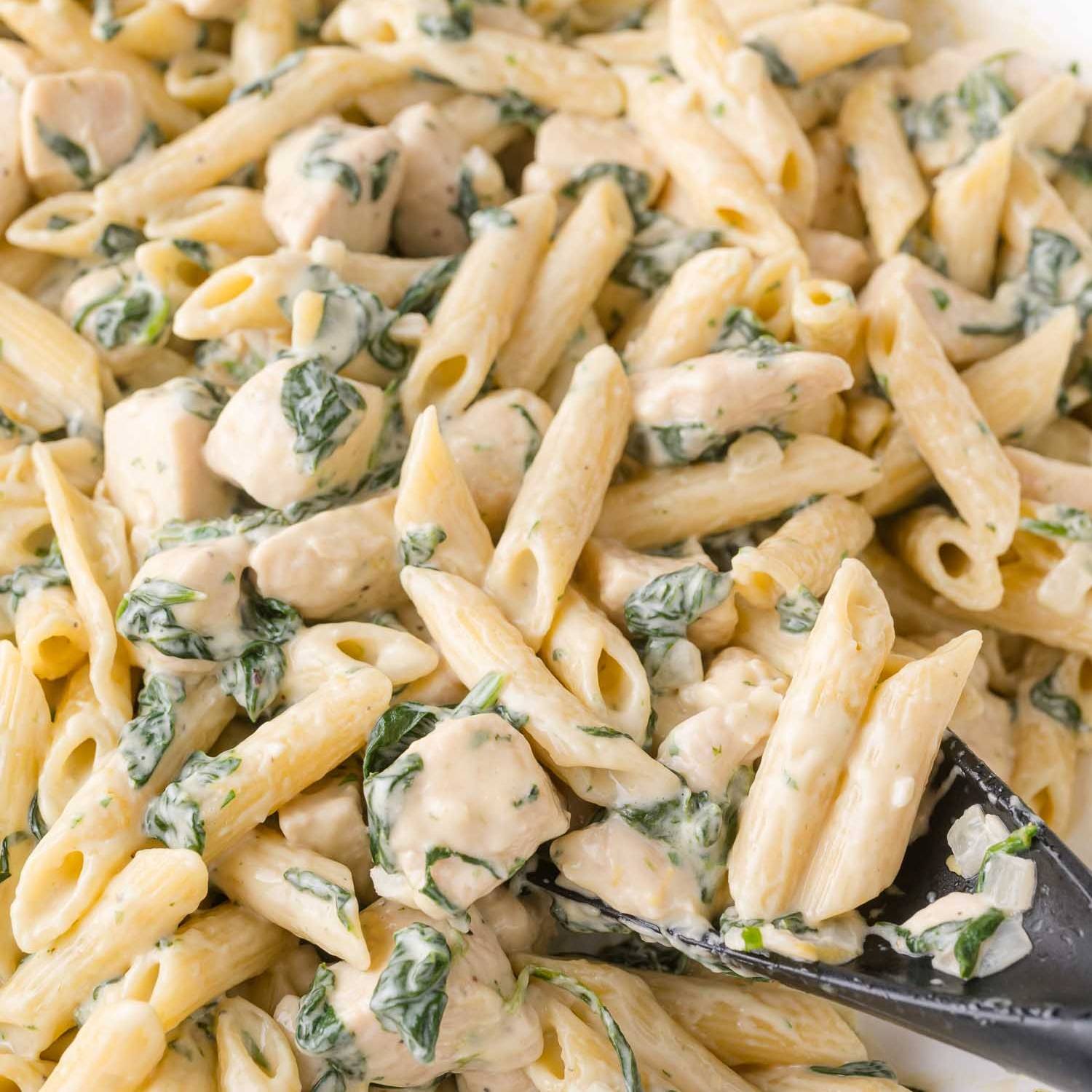 Penne pasta in a creamy sauce with cooked chicken and spinach. A spoon scoops some of it up.