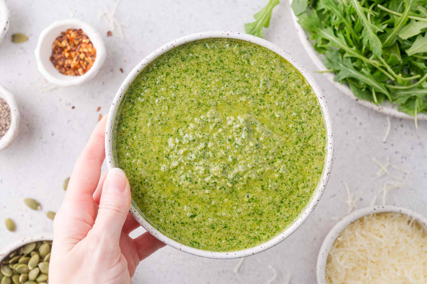 Arugula pesto in a bowl, held in a hand, with ingredients in the background (arugula, cheese, red pepper flakes, pepitas).