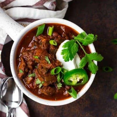 Dark red chili in a white bowl topped with sour cream, green onion, cilantro.