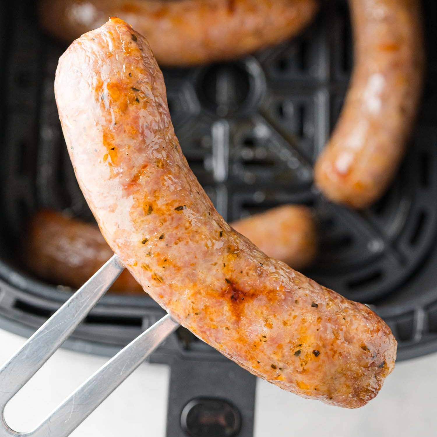 Air fryer Italian sausage held with a fork over an air fryer with more sausages.