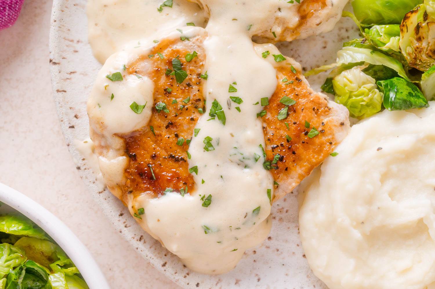 Pork chop with creamy garlic sauce on a plate with potatoes and Brussels sprouts.