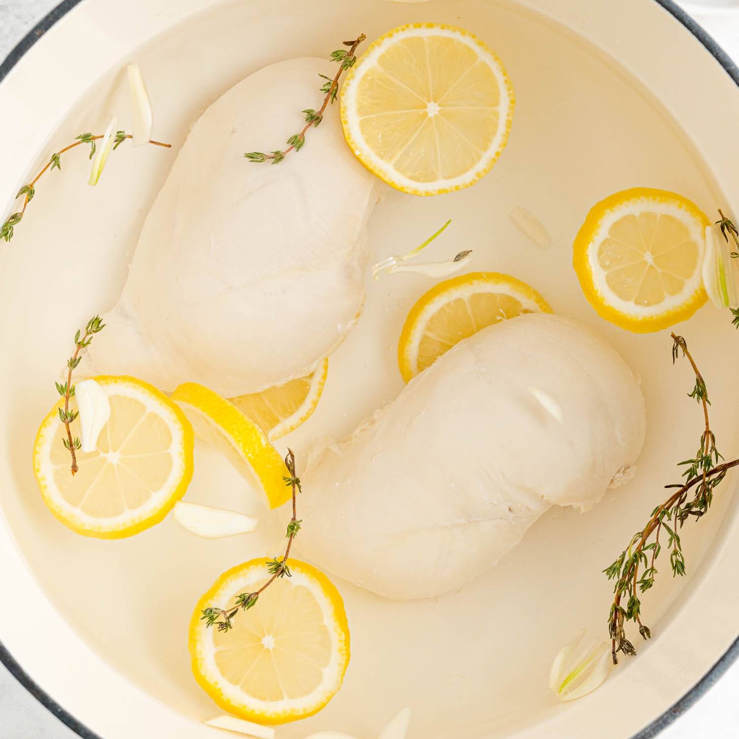 Two poached chicken breasts in a pan with lemon slices and thyme sprigs.