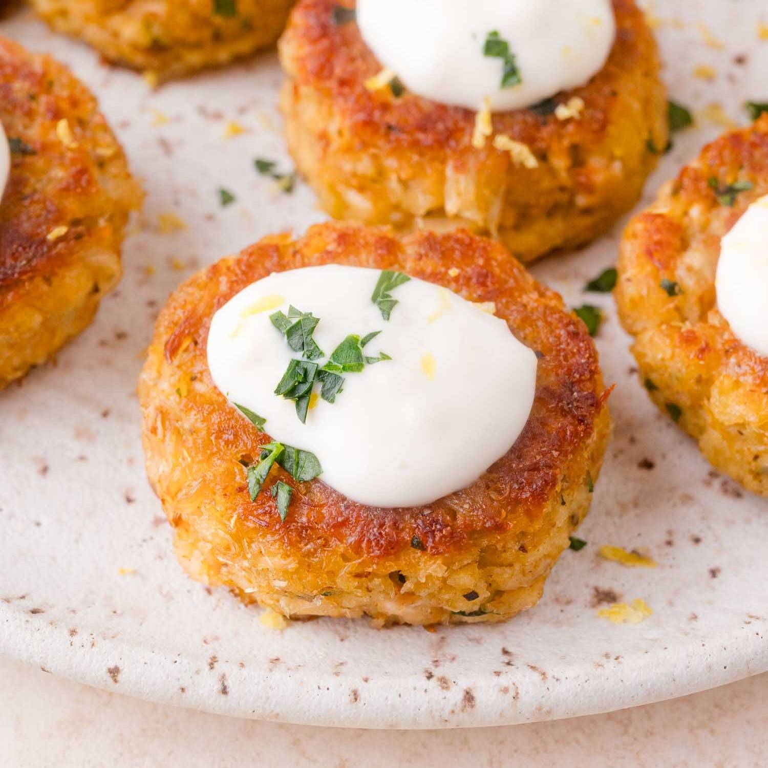 Mini crab cakes on a plate, topped with aioli and fresh herbs.