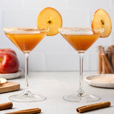 Two martini galsses with apple cider in them, garnished with a thin sliced apple.