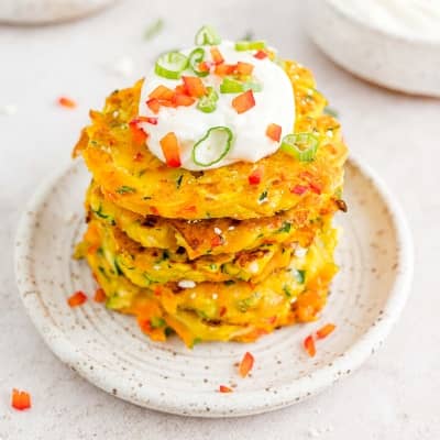 Stack of vegetable fritters topped with sour cream and chopped red bell peppers.