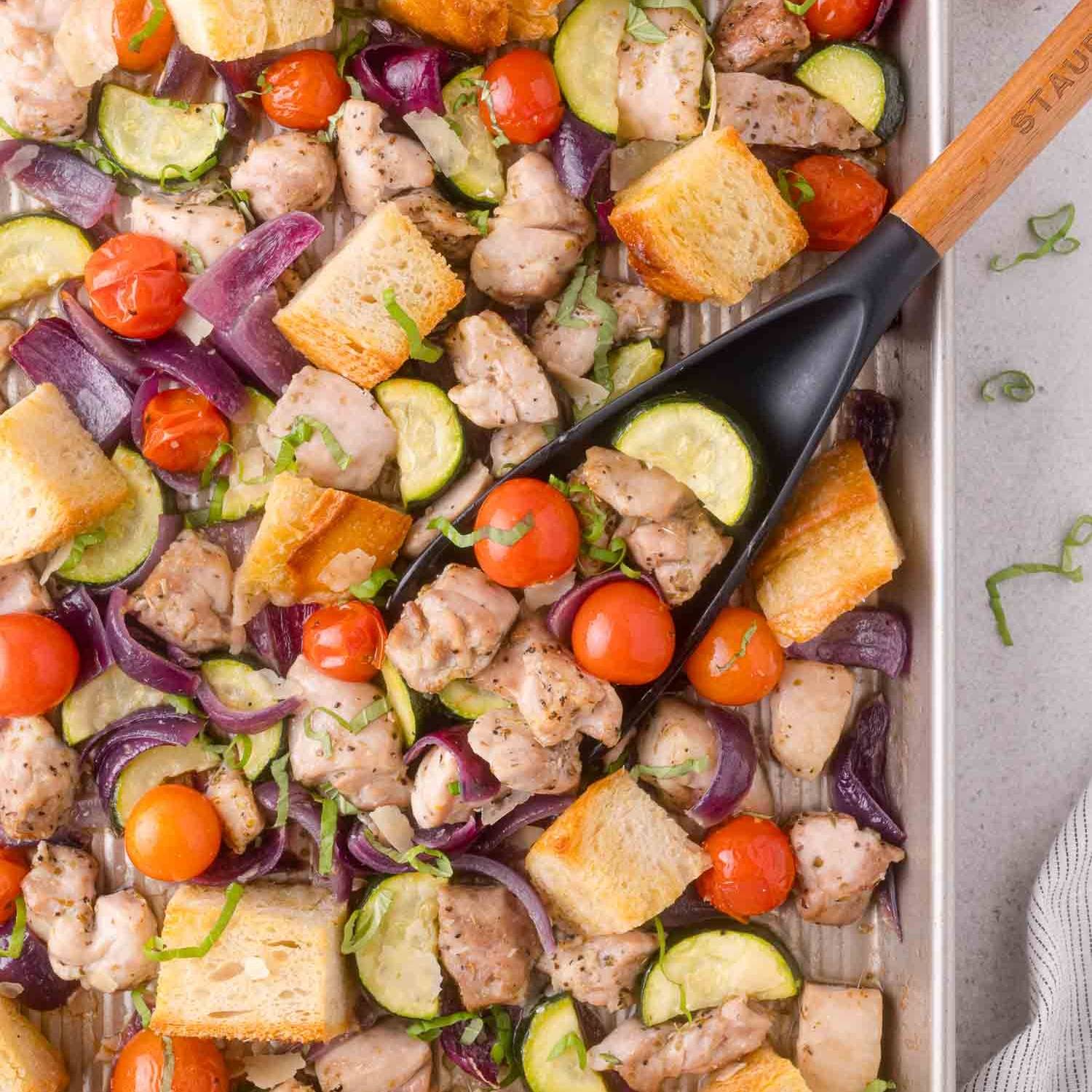 Sheet pan with chicken, tomatoes, red onion, zucchini, and bread cubes.