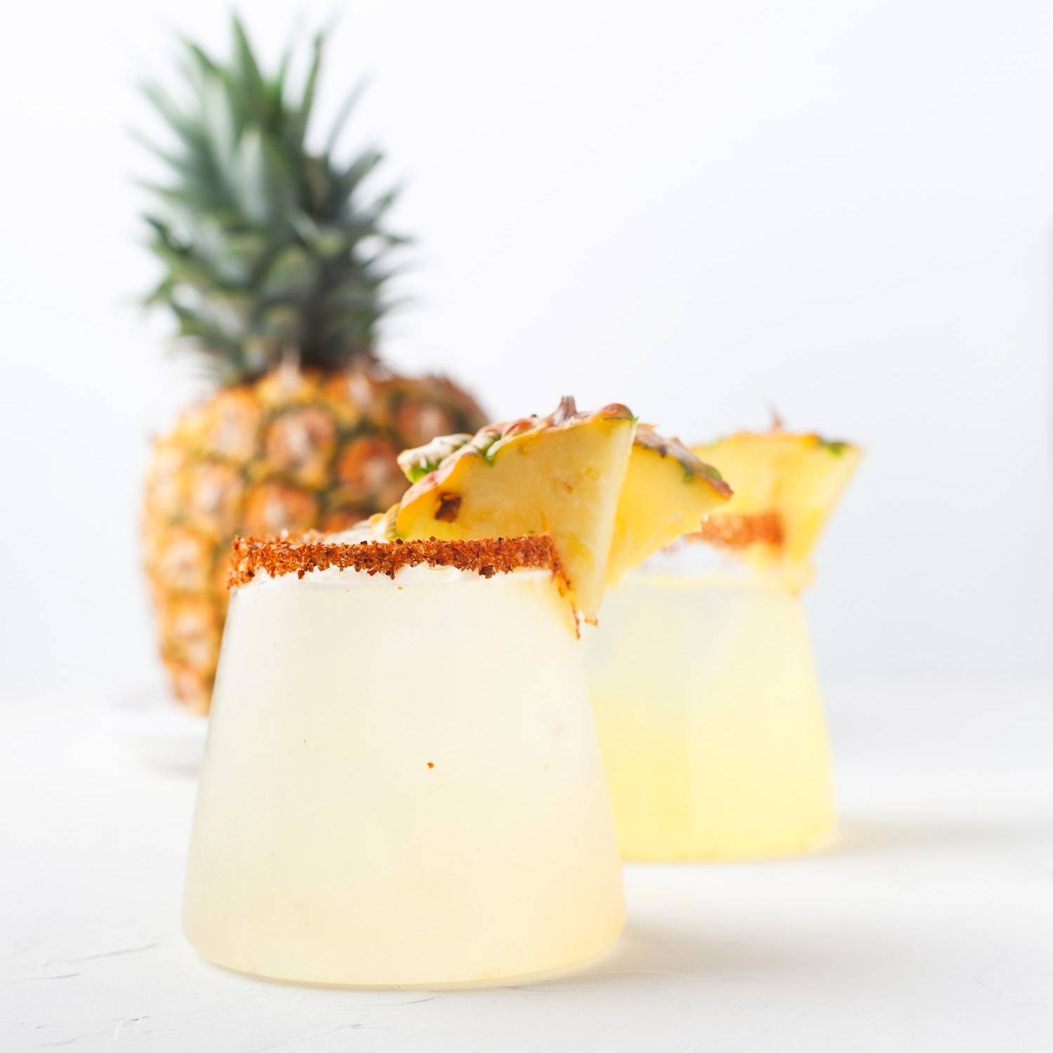 Pineapple margarita with a spice and salt rim, garnished with fresh pineapple, another one in the background.
