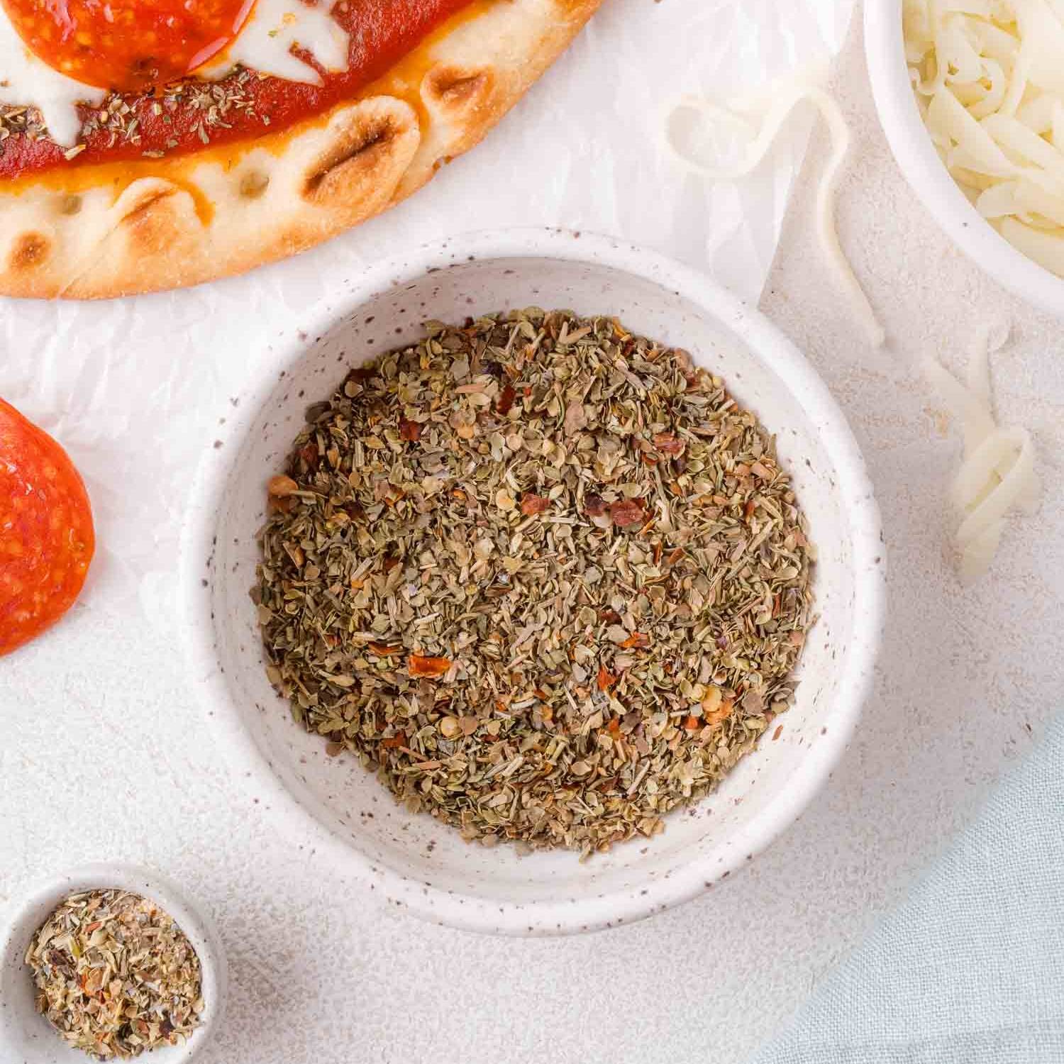 Pizza seasoning in a small bowl, with a pizza partially visible in the corner.