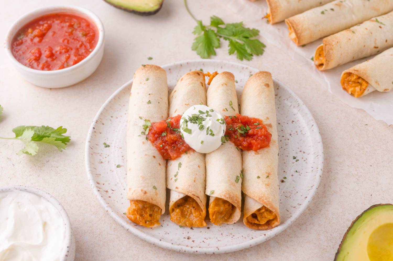 Four baked chicken taquitos on a plate topped with sour cream and salsa. 